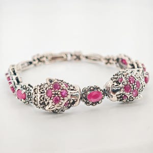 May include: Silver-toned bracelet featuring panther head designs and oval-shaped ruby-colored gemstones. The bracelet has a detailed, ornate design with multiple links and a secure clasp. The gemstones are set within the panther heads and along the bracelet.