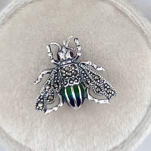 Puede incluir: Un broche de abeja de plata con un cuerpo de esmalte verde y negro y detalles de marcasita. El broche se muestra en un cojín de terciopelo blanco.