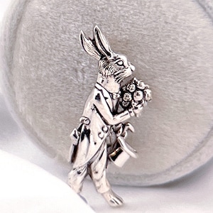 Może przedstawiać: Srebrny charm przedstawiający królika w garniturze trzymającego bukiet kwiatów. Charm znajduje się na białym tle.