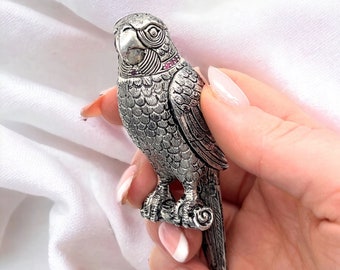 Puede incluir: Una figurita de loro de plata con detalles en rosa. El loro está posado en una rama y tiene plumas detalladas.