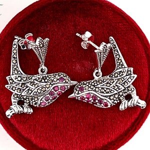 May include: A pair of silver earrings with a bird design. The birds are facing each other and have pink gemstones on their bodies. The earrings are on a red velvet background.