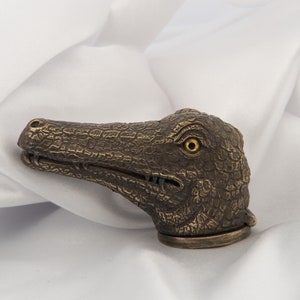 May include: A bronze alligator head bottle stopper with a textured surface and a yellow eye. The stopper is on a white background.