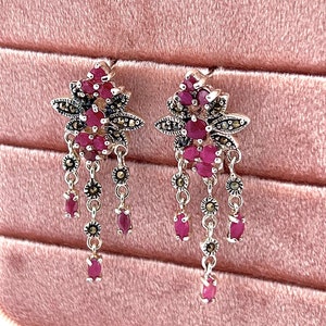 以下が含まれることがあります： 赤いルビーと黒いマルカジットのアクセントが付いたシルバーのドロップイヤリングのペア。イヤリングは、花の形をしたルビーとマルカジットのクラスターを特徴とし、ルビーとマルカジットの3本の垂れ下がったチェーンが付いています。