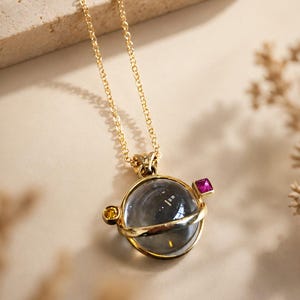 Puede incluir: Collar dorado con un colgante esférico. El colgante tiene un orbe transparente y grisáceo, una banda dorada y está adornado con una gema amarilla y otra rosa. La fina cadena dorada es adecuada para el uso diario. Una joya.