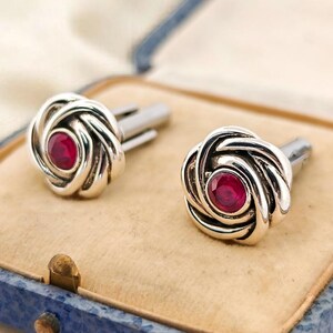 May include: A pair of silver cufflinks with a rose design, each featuring a round, red gemstone center. The cufflinks are displayed in a vintage blue box with a beige interior, set against a cream-colored fabric.