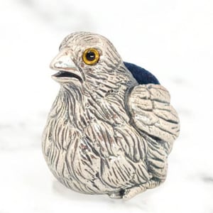 Puede incluir: Un cojín para alfileres con forma de pájaro de color plateado con una inserción de tela azul. El pájaro tiene texturas de plumas detalladas, un ojo amarillo y un pico ligeramente abierto. El objeto está colocado sobre una superficie de mármol blanco.