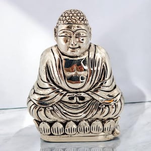 May include: A silver-colored Buddha statue in a seated meditation pose. The statue has a serene expression and is detailed with intricate folds in the robes and a textured head. The base is a lotus flower design.
