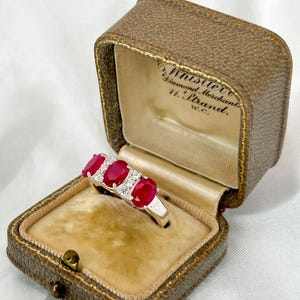 May include: A ring with three red ruby stones and smaller white diamonds set in a silver band. The ring is in a brown velvet ring box with the inscription "Whistler Diamond Merchant H. Brand, W.C."