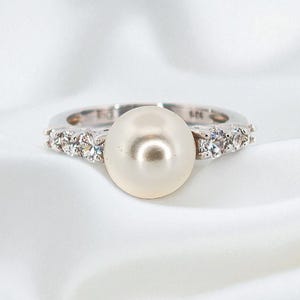 May include: A silver ring featuring a large, lustrous pearl as the centerpiece. The band is adorned with small, sparkling gemstones. The ring is set against a white, silky background, highlighting its elegant design.