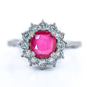 Può includere: Anello in argento con una grande gemma rosa vivace al centro, circondata da gemme azzurre più piccole. La fascia dell'anello presenta un design strutturato e organico.