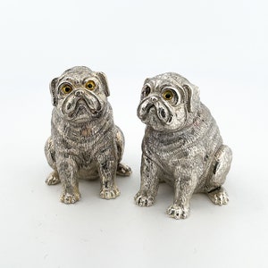 May include: Two silver pug figurines with yellow eyes. The pugs are sitting and facing forward.