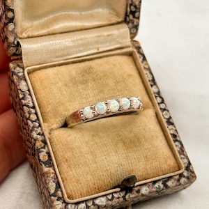 May include: A silver ring featuring five opal gemstones, presented in a vintage jewellery box. The opals are circular and set in a line. The box has a beige fabric lining and a patterned exterior.