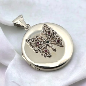 May include: Silver locket featuring a filigree butterfly design. The butterfly is adorned with red and green gemstones.