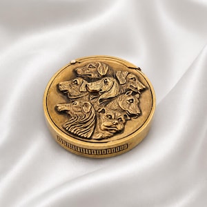 May include: A round, gold-toned compact featuring a bas-relief design of several dog breeds. The dogs are intricately detailed, with visible fur and facial features. The compact is set against a white, silky background.