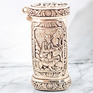 Puede incluir: Un recipiente de metal beige con un detallado relieve de un cazador con un perro. El recipiente tiene una tapa con bisagras y patrones florales decorativos en la parte superior e inferior. El recipiente es de forma cilíndrica.