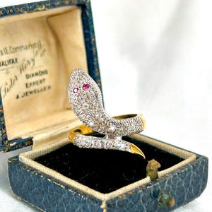 May include: A gold ring with a diamond-encrusted snake design. The snake's eyes are ruby red. The ring is in a blue velvet box with the text "Lister & Co. Halifax Diamond Expert & Jewellers" on the lid.