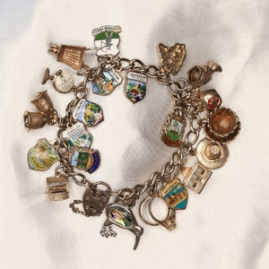 Puede incluir: Un brazalete de plata con una variedad de dijes, incluyendo un pájaro, una casa, una campana y un corazón. Los dijes son todos de plata y tienen diferentes detalles de esmalte de colores. El brazalete está sobre un fondo blanco.