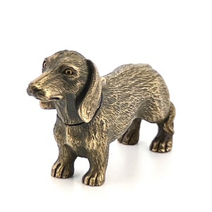 May include: A small, bronze-colored dachshund figurine. The figurine is standing on all four legs and has a detailed, realistic appearance.