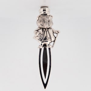 Puede incluir: Marcapáginas de plata con una pequeña figura detallada de un ratón con un sombrero de copa y un bastón. El marcapáginas tiene forma de hoja larga y fina.