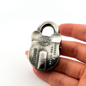 May include: A silver-toned metal padlock featuring the engraved text CHUBB LONDON, MAKERS TO THE QUEEN, and the serial number 1235526. The padlock has a rounded body with a prominent central vertical plate and a curved shackle at the top.