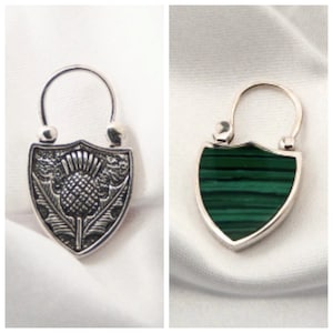 May include: Two shield-shaped pendants. One is silver with a thistle design, the other has a green malachite stone. Both have a silver bail at the top. The thistle pendant has the number "925" engraved on it.