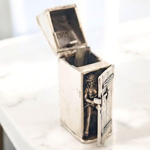 May include: A silver-toned, rectangular box with an open lid. The box features a small, detailed figure inside, with a door ajar. The box is set on a white marble surface.