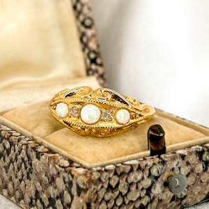 Peut inclure: Une bague en or avec trois opales blanches et de petits diamants sertis dans un design délicat et orné. La bague est présentée dans un écrin à bague en velours brun.