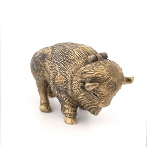 May include: A small, gold-coloured metal sculpture of a bison. The bison is standing with its head lowered and its horns visible. The sculpture is detailed with textured fur and a hinged back.