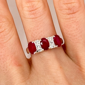 May include: A silver ring with three oval-shaped red gemstones and smaller clear gemstones set in the band.