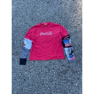 Puede incluir: Camiseta roja de manga larga con el logotipo de Coca-Cola en la parte delantera. Las mangas están hechas de diferentes telas con varios diseños gráficos, incluida una bandera estadounidense y un águila.