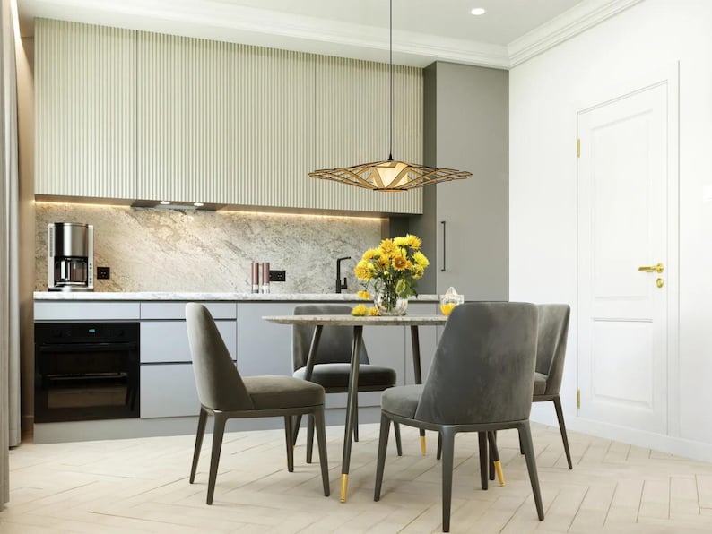 Op de afbeelding: Een moderne eetkamer met een lichte houten vloer, een witte keuken met grijze kasten, een zwarte oven en een tafel met vier grijze stoelen. Een geometrische lamp hangt boven de tafel. Een boeket gele bloemen staat op de tafel.