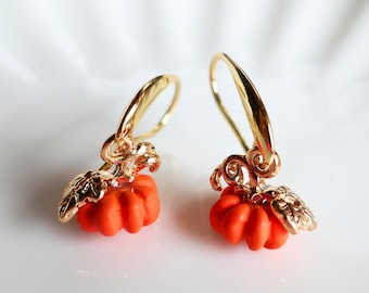 May include: A pair of gold-tone drop earrings featuring small, vibrant orange pumpkin charms. Each pumpkin has a textured gold-tone stem and leaf detail attached to a curved gold-tone ear wire. The text Beverly Mint Designs is visible.
