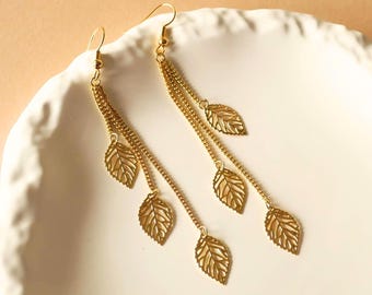 May include: A pair of gold-toned drop earrings featuring multiple delicate chains suspended from a hook. Each chain is adorned with small, intricate, cut-out leaf-shaped charms. The design creates a layered, cascading effect with a polished finish.