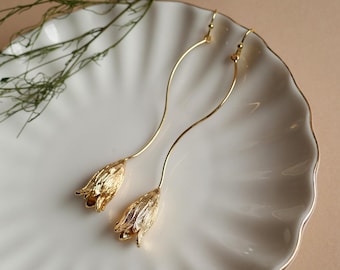 May include: A pair of long gold-toned earrings featuring a slender, curved wire stem that leads to a textured, bell-shaped flower pendant. The earrings hang from simple hook closures, showcasing a metallic finish with intricate petal detailing.