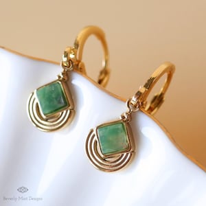 May include: A pair of gold-toned earrings with a square, light green gemstone center. The earrings have a geometric design with a half-circle pattern below the gemstone. The earrings are displayed on a white surface.