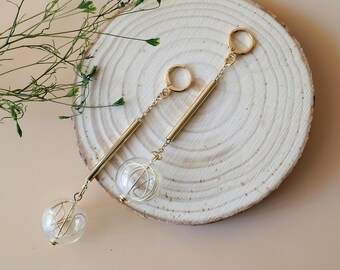 May include: A pair of gold-toned dangle earrings featuring a circular hoop top, a thin metallic bar, and a clear glass sphere containing a delicate gold wire coil. The earrings are asymmetrical in length, showcasing a refined metallic design.