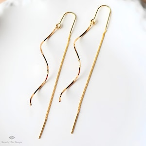 May include: A pair of gold-colored threader earrings. Each earring features a curved, wavy design and a delicate chain that hangs down. The earrings are displayed on a white surface.