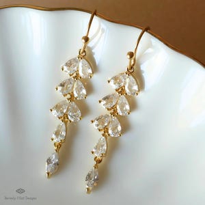 Peut inclure: Boucles d'oreilles pendantes dorées avec un design en cascade de cristaux transparents en forme de poire et de pétales. Les boucles d'oreilles sont présentées sur une surface blanche, mettant en valeur leur éclat et leurs détails. Un accessoire délicat.