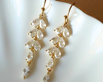 May include: A pair of gold-toned drop earrings featuring a cascading vine design. Each earring is composed of multiple clear, pear-shaped and marquise-cut crystals arranged in a vertical, leaf-like pattern, suspended from a simple gold hook wire.