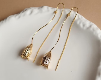 May include: A pair of gold-tone threader earrings featuring a textured, bell-shaped flower pendant at the end of a curved wire. The earrings have a long, slender chain design that hangs down, with a polished metallic finish and a delicate floral motif.