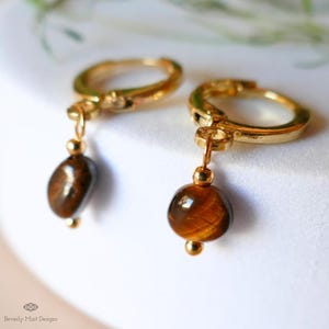 May include: Gold-plated hoop earrings, each featuring a polished, brown tiger's eye gemstone. The earrings have a classic design, with a small, round bead above the gemstone. A simple, elegant jewellery item.