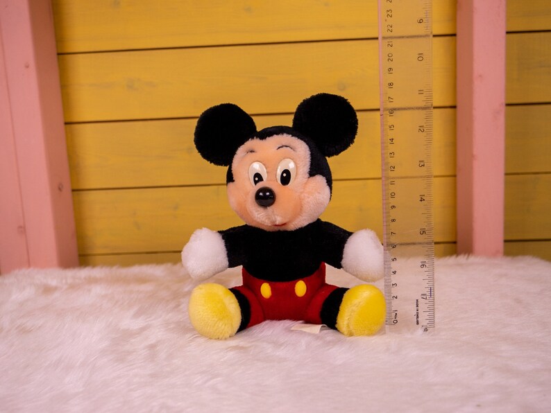 以下が含まれることがあります： 黒と赤のぬいぐるみミッキーマウスの玩具。大きな黒い耳と白い顔をしています。玩具は白いふわふわの表面の上に座っています。