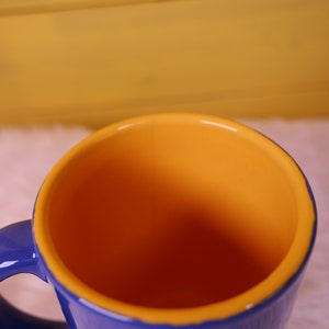 May include: A blue ceramic mug with a yellow interior. The mug has a handle and is empty.