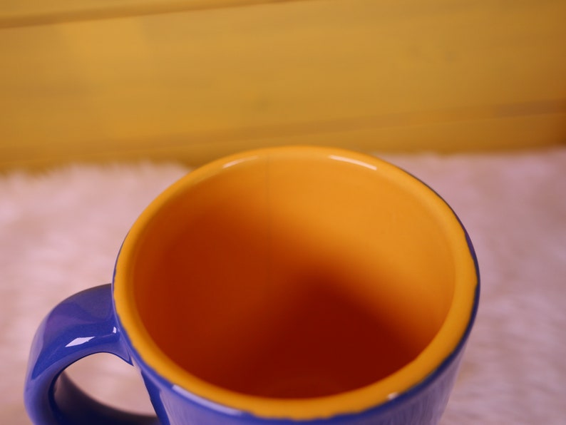 May include: A blue ceramic mug with a yellow interior. The mug has a handle and is empty.