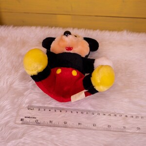 以下が含まれることがあります： 黒い耳、赤と黒の体、黄色い手と足をつけたミッキーマウスのぬいぐるみ。おもちゃは白いふわふわの表面に置かれています。