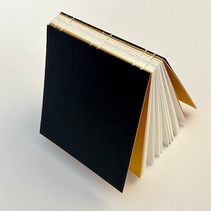 Op de afbeelding: Een handgemaakt notitieboek met een zwarte kaft en goudkleurige binnenpagina's. De pagina's zijn wit en gebonden met witte draad. Het notitieboek is open, wat het contrast tussen de zwarte kaft en de gouden en witte pagina's laat zien.