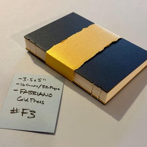 Pode incluir: Um diário feito à mão com capa azul marinho e faixa dourada. O diário mede aproximadamente 8,9 x 12,7 cm e contém 16 folhas/32 páginas de papel Fabriano prensado a frio. Um pequeno cartão azul exibe os detalhes e o código #F3.