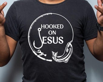 Könnte beinhalten: Ein schwarzes T-Shirt mit einer weißen Grafik eines Angelhakens und eines Fisches. Die Grafik sagt "Hooked on Jesus".