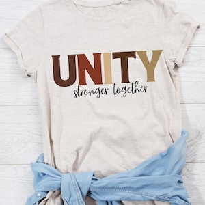 May include: A beige t-shirt with the word "UNITY" in brown, tan, and gold letters. Below the word "UNITY" is the phrase "stronger together" in black script. A light blue denim jacket is tied around the waist of the shirt.