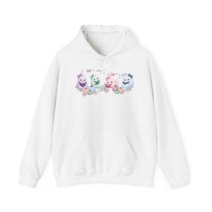 Peut inclure: Un sweat à capuche blanc avec un motif central représentant quatre fantômes aux couleurs pastel portant des nœuds et lisant des livres. Les fantômes sont entourés de petites citrouilles, de feuilles d'automne et d'étoiles violettes, vertes, roses et bleues.